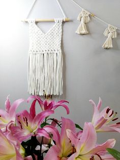 pink lilies are in a vase next to a wall hanging with tassels