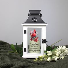 a small white lantern with a cardinal on it and flowers in the corner next to it