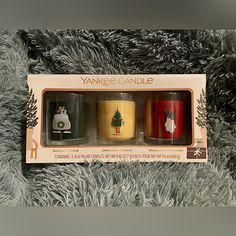 three yankee candles are sitting in a box on a fur covered surface, one is yellow and the other is red