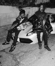 two young men sitting on the hood of a sports car in front of a wall