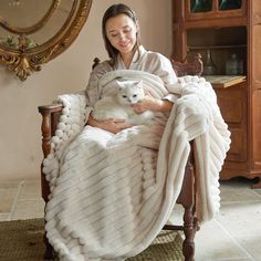 a woman sitting in a chair holding a white dog under a blanket on her lap