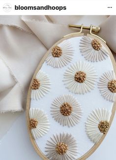 an embroidery project with white flowers and gold accents on a wooden hoop hanging from a satin fabric