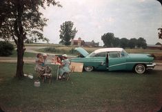 two women sitting at a table in front of an old car with the hood open