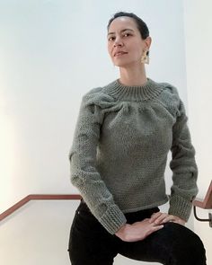 a woman sitting on top of a chair wearing a green sweater and black pants with her hands in her pockets