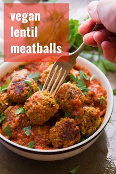 a hand holding a fork over a bowl of meatballs with sauce and parsley