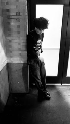 a young man sitting on the side of a building next to a door with his hands in his pockets