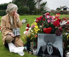 an elderly woman sitting on the ground in front of flowers and a memorial with a book