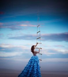 a woman in a blue dress standing on top of a ladder looking up at the sky