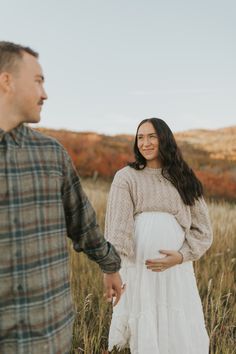 a pregnant woman holding her husband's hand while they walk through the tall grass