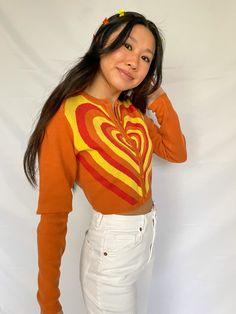 cropped orange cardigan with red, orange, and yellow heart design available in sizes xs, s, m, l model is 5'3" and wearing a size small Orange Retro Long Sleeve Sweater, Retro Orange Long Sleeve Sweater, Orange Long Sleeve Retro Sweater, Fitted Heart Print Sweater For Fall, Orange Fitted Long Sleeve Cardigan, Fitted Orange Long Sleeve Cardigan, Fitted Long Sleeve Orange Cardigan, Trendy Orange Crop Top For Fall, Casual Orange Crop Top For Fall