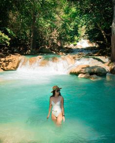 a woman in a white bathing suit and hat standing in the water near a waterfall