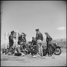 black and white photograph of men standing around motorcycles on the side of the road with one man laying down