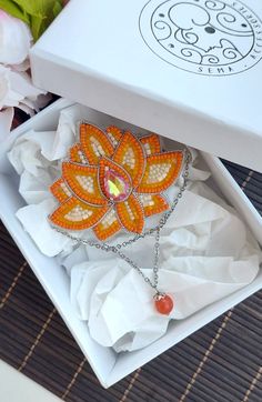 an orange and yellow brooch sits in a white box next to some pink flowers