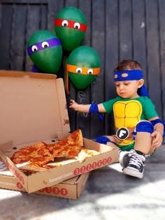 a young boy sitting on the ground next to a box of pizza and some balloons