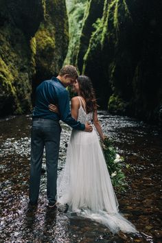 a bride and groom standing in the middle of a stream with mossy cliffs behind them