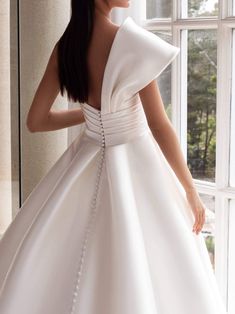 a woman in a white wedding dress standing by a window with her back turned to the camera