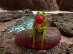 a toy frog sitting on top of a rock with a red ball in its mouth