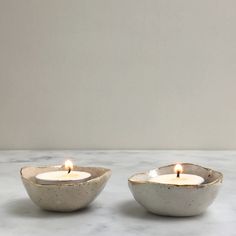 two white ceramic bowls with lit candles in them on a marble countertop next to each other