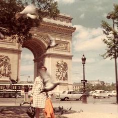 a woman standing in front of a large arch with birds flying around her and cars parked on the street