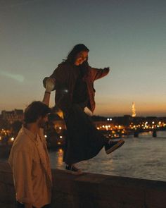 a man and woman standing on the edge of a bridge over water at night with city lights in the background