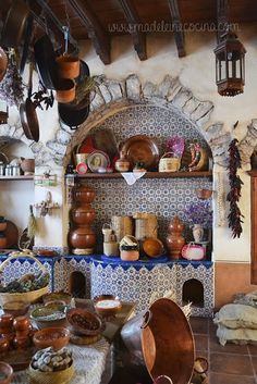 an old kitchen with pots and pans hanging from the ceiling, in front of a stone fireplace