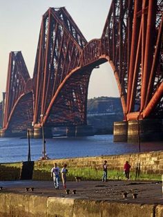 some people are walking on the sidewalk by the water and an old bridge is in the background