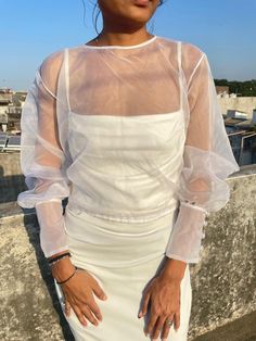 a woman wearing a white dress standing on top of a roof with her hands in her pockets