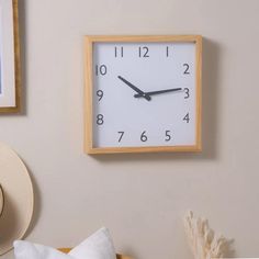 a square clock mounted to the side of a wall next to a hat on a bed