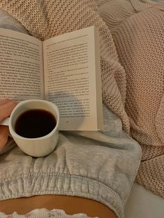 a person is reading a book and holding a cup of coffee while laying in bed