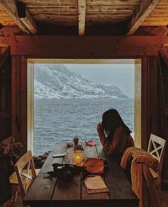 a woman sitting at a table looking out over the ocean from inside a wooden cabin