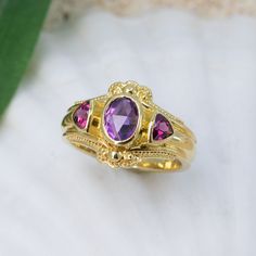 Adorn yourself with this stunning, 18K granulated golden ring showcasing a majestic combination of beautiful amethyst and vibrant rhodolite garnets. Add a regal touch to any occasion and feel like a queen. 18KY granulated ring with 7x5mm uniquely-cut amethyst and 2- 4mm rhodolite garnets. The granulation on runs part way down the band resulting in a band width of 4.7mm. Currently size 6.75, but each ring comes with free sizing and FREE SHIPPING! Unsure what ring size you are? It's always best to Exquisite Yellow Gold Amethyst Ring With Accent Stones, Gold Garnet Birthstone Ring, Heirloom Multi-stone Amethyst Ring, Yellow Gold Rings With Pink Sapphire And Gemstone Accents, Luxury Amethyst Rings With Rose Cut Diamonds, Gold Sapphire Ring With Accent Stones, Gold Heirloom Ruby Ring With Gemstone Accents, Gold Amethyst Rings With Rose Cut Diamonds, Gold Sapphire Ring With Pink Sapphire Accent Stones