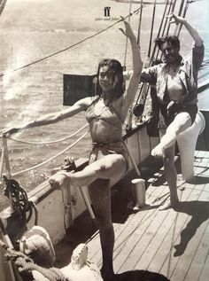 two women in bathing suits standing on the deck of a sailboat with their arms stretched out