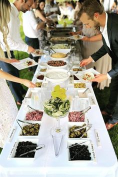 several people are serving themselves food at an outdoor buffet line up on the grass with white tablecloths