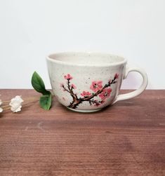 a white cup with pink flowers on it sitting on a wooden table next to a flower