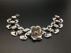 "This pretty bracelet is made of very lightweight silver-tone metal.  The center focal point is a raised rose with a rhinestone center.  Either side of the rose are silver leaves with small flowers with rhinestone centers.  It has a circular slide lock closure.  It measures 6 1/2\" long and about 1\" wide and is in beautiful condition as shown." Silver Metal Jewelry With Rose Design, Silver Flower Shaped Jewelry With Rose Design, Silver Floral Jewelry With Rose Design, Silver Flower Jewelry With Rose Design, Silver Rose Design Jewelry For Formal Occasions, Elegant Silver Bracelet With Rose Design, Elegant Silver Bracelets With Rose Design, Silver Leaves, Pink Milk