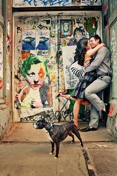 a man and woman kissing in front of a graffiti covered building with a dog on a leash