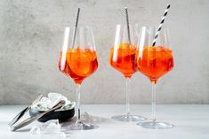 three wine glasses filled with orange liquid next to ice cubes and silverware on a white surface