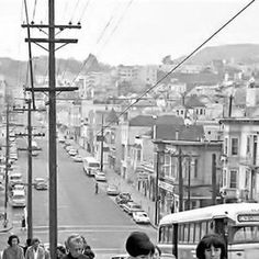 black and white photograph of people walking down the street in front of cars, buses and telephone poles