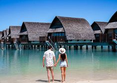 a man and woman holding hands while standing on the beach in front of some huts