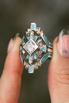 A close-up image of a woman's fingers holding a "Calliandra" design three-piece wedding ring set from Staghead Designs. This ring set features geometric Western-style design elements & a 2ct princess cut moissanite engagement ring with crushed turquoise inlays & diamond accents & is paired with two matching turquoise & diamond bands. This ring is shown in 14K white gold but can also be made in yellow or rose gold. Western Wedding Ring Stacker, Engagement Rings With Unique Stones, Navajo Wedding Rings, Unique Turquoise Rings, Vintage Ring Sets, Turqouise Wedding Ring, Southwestern Engagement Ring, Native Wedding Rings, Southwest Engagement Ring