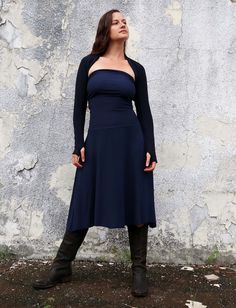 a woman standing in front of a concrete wall wearing boots and a dress with long sleeves