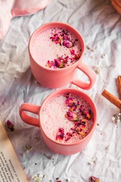 two pink mugs filled with liquid and flowers next to an open book on a table