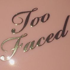 the word too faced is written in cursive writing on a pink surface with silver foil