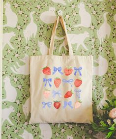 a tote bag with strawberries and bows on it hanging from a wall next to flowers