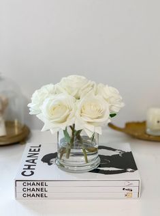 white roses in a glass vase sitting on top of a stack of chanel books