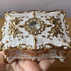 a hand holding a white and gold box with an ornate design on it's side