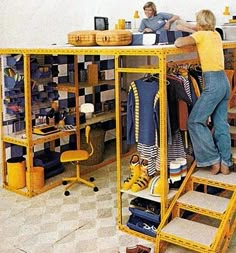 two people standing in front of a yellow shelf filled with clothes and other items on it