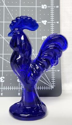 a blue glass rooster figurine sitting on top of a table next to a ruler