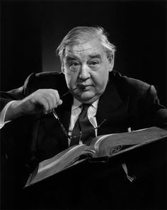 an old man sitting in a chair holding a book and looking at the camera while reading it