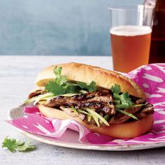 a sandwich with meat, lettuce and cilantro on a plate next to a glass of beer